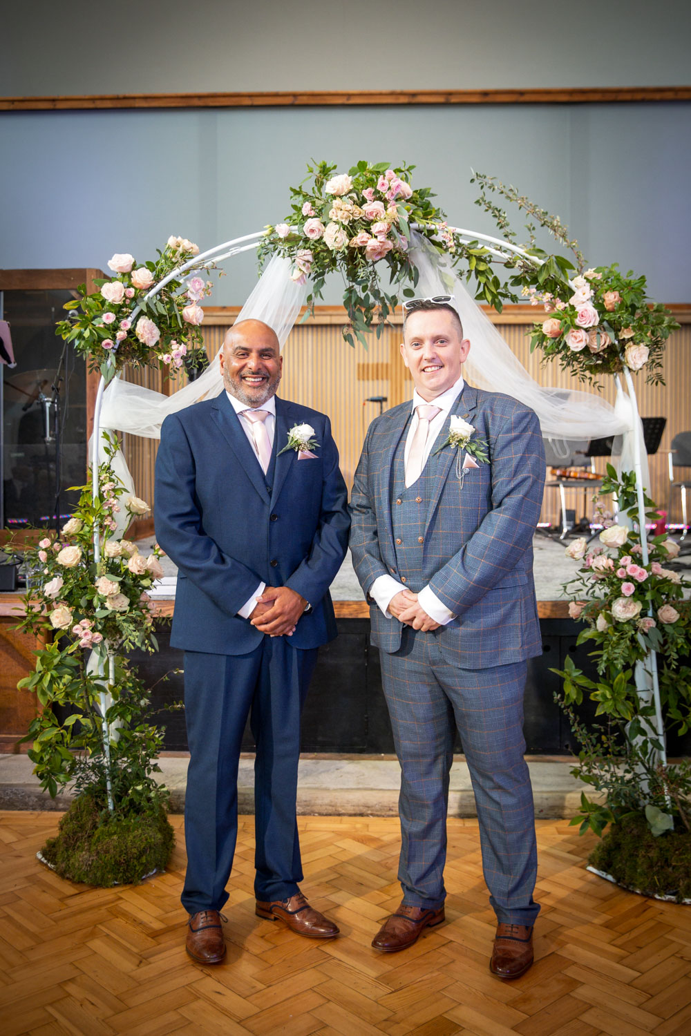 Two grooms smiling under floral arch indoor wedding by Southampton wedding photographer John Watson