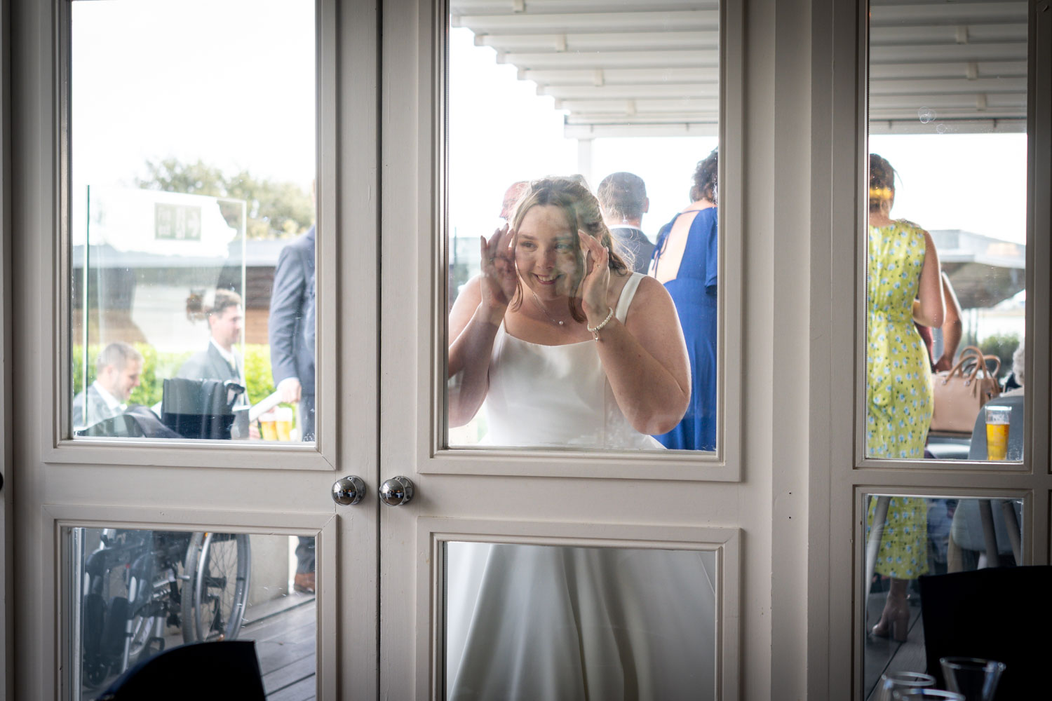 Bride making funny faces through glass door by Southampton wedding photographer John Watson