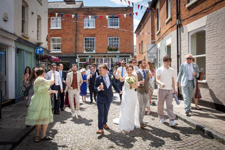 Bride and groom walk with guests on sunny street by Southampton wedding photographer John Watson